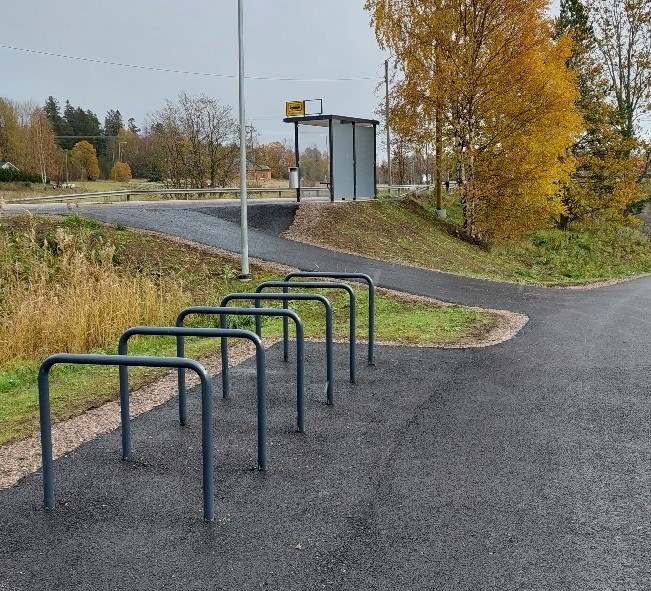 Pöyräpysäköintipaikka linja-autoasemalla. Metalliset kaaret etualalla. Taustalla linja-autopysäkki. Harmaa taivas, taustalla puita, joiden lehdet ovat kellastuneet.