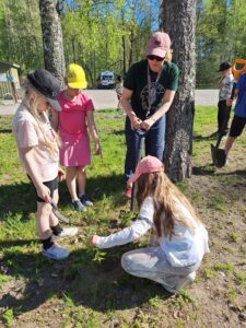 Kaksi tyttöä ja opettaja katselevat, kun yksi tyttö on kyykistyneenä maahan ilmeisesti tarkastelemaan kasvia. Opettajalla on kädessään iso lapio. Tarkoituksena on poistaa kasvit uuden niittykasvillisuuden tieltä.